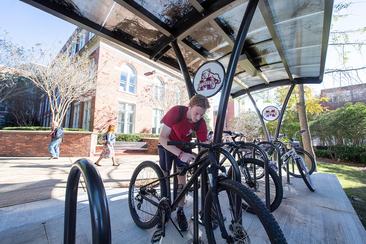 A student is securing a bicycle at a bike rack under a covered shelter on a sunny day.